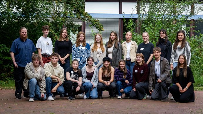 Das Bild zeigt 19 der 22 Freiwilligen, die sich zu einem Gruppenbild aufgestellt haben. Sie stehen vor einem Unigebäude und lächeln in die Kamera.