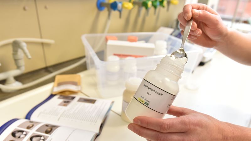 Eine Hand hält ein durchsichtiges PLastikfläschchen, auf dem Ammoniumnitrat steht. Die andere Hand hält einen Löffel, mit dem sie ein Pulver aus der Flasche entnimmt.