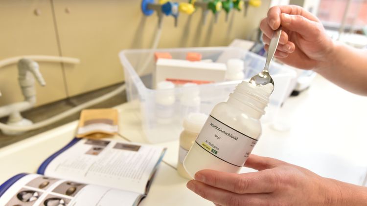 Eine Hand hält ein durchsichtiges PLastikfläschchen, auf dem Ammoniumnitrat steht. Die andere Hand hält einen Löffel, mit dem sie ein Pulver aus der Flasche entnimmt.