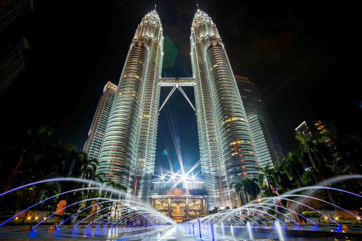 Das Bild zeigt die Petronas Twin Towers in Kuala Lumpur, Malaysia, bei Nacht. Die Türme sind beleuchtet und ragen majestätisch in den dunklen Himmel. Vor den Türmen befindet sich ein Brunnen mit Wasserfontänen, die durch Lichteffekte hervorgehoben werden. Die Szene vermittelt einen Eindruck von moderner Architektur und urbaner Pracht.