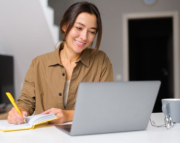 Freundliche junge Frau arbeitet von zu Hause aus. Sie sitzt am Küchentisch, vor dem Laptop und dem Notizbuch. 