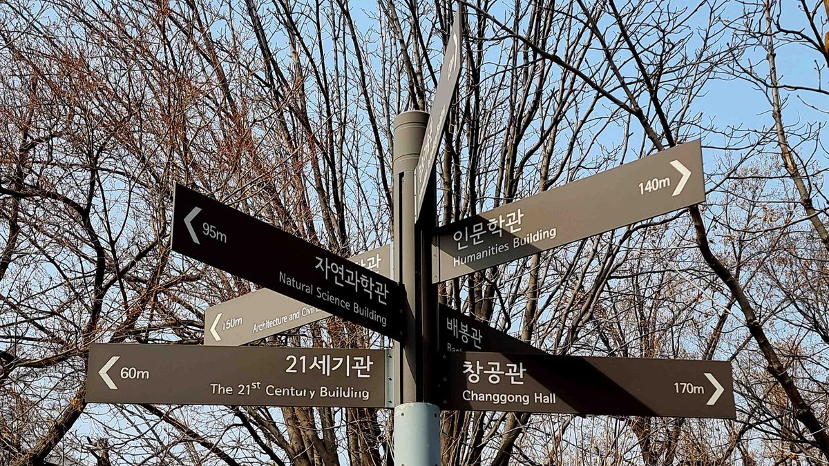 Das Bild zeigt einen metallenen Wegweiser auf dem Campus der University of Seoul. Der Wegweiser ist mit mehreren Schildern in koreanischer und englischer Sprache versehen, die verschiedene Gebäude und Entfernungen anzeigen, darunter das "Humanities Building", das "Natural Science Building" und den "Changgong Hall". Im Hintergrund sind kahle Bäume mit filigranen Ästen zu sehen, die den Himmel durchziehen