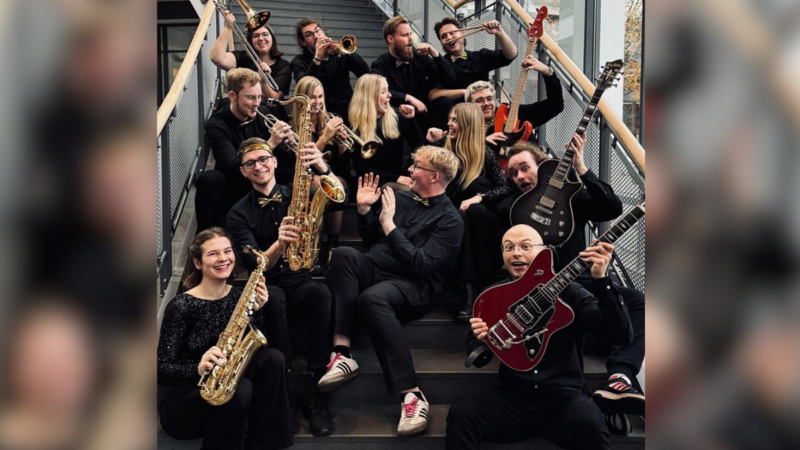 Das Bild zeigt die 14 Mitglieder von „Funkystition“. Sie sitzen auf einer Treppe im Hörsaalgebäude A 14 und lachen und freuen sich. Sie halten ihre Instrumente in den Händen, darunter Blasinstrumente und Gitarren.