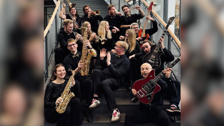 Das Bild zeigt die 14 Mitglieder von „Funkystition“. Sie sitzen auf einer Treppe im Hörsaalgebäude A 14 und lachen und freuen sich. Sie halten ihre Instrumente in den Händen, darunter Blasinstrumente und Gitarren.
