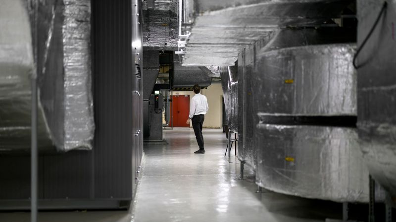 Blick in die Technikzentrale, man sieht große, mit metallisch glänzemden Dämmmaterial umwickelte Luftkanäle. Dazwischen schaut ein Mann, der nach oben schaut und zwischen den gewaltigen Kanälen eher klein wirkt.  