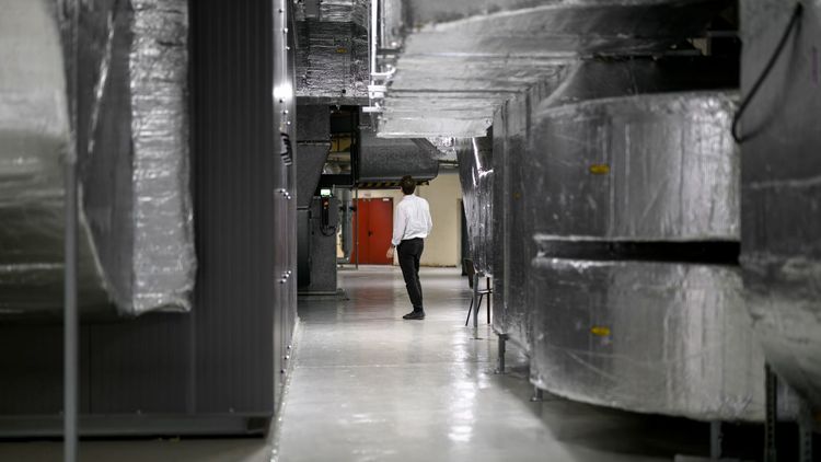 Blick in die Technikzentrale, man sieht große, mit metallisch glänzemden Dämmmaterial umwickelte Luftkanäle. Dazwischen schaut ein Mann, der nach oben schaut und zwischen den gewaltigen Kanälen eher klein wirkt.  