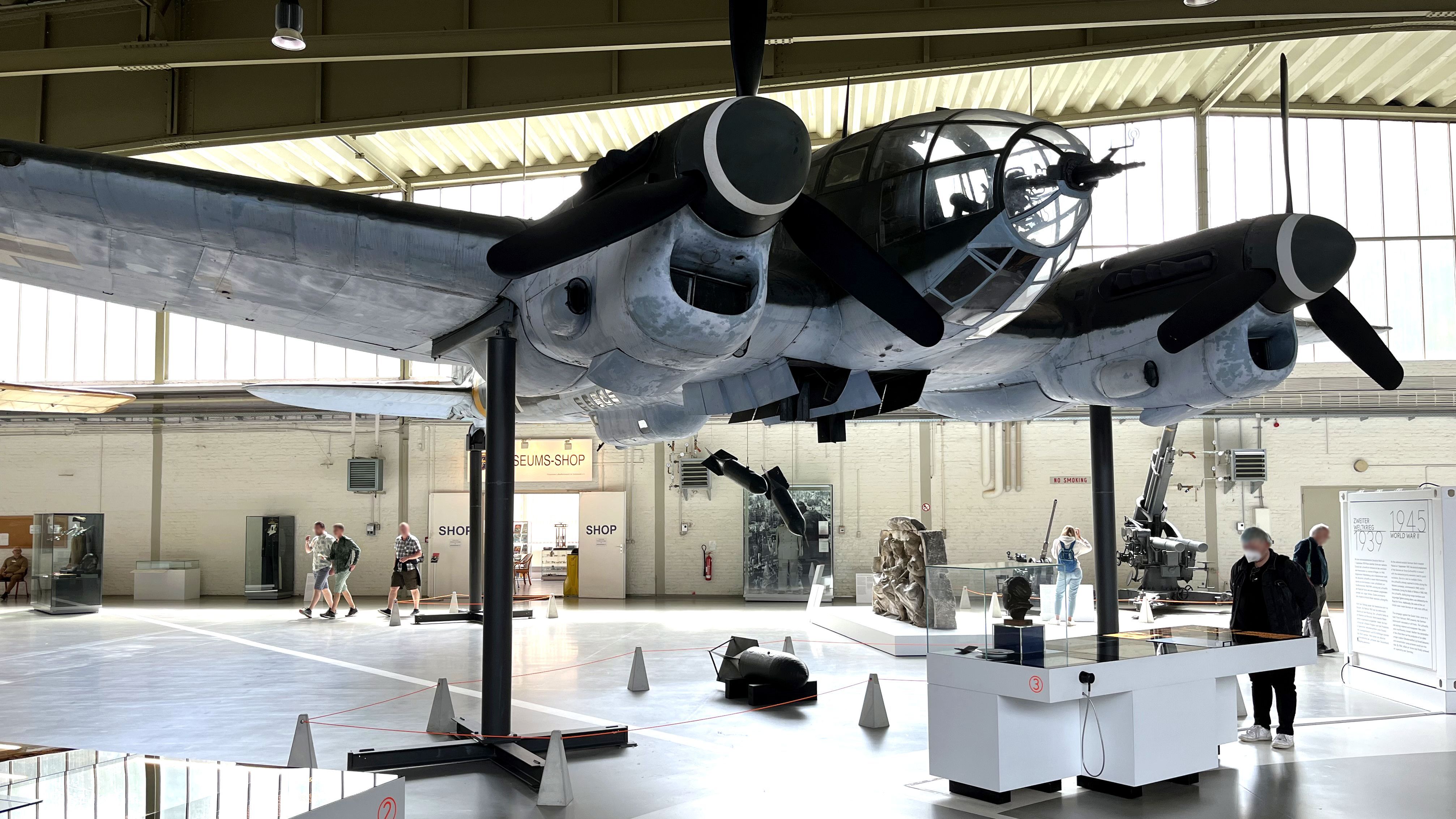 The picture shows a model of a bomber in a large museum hall. The aircraft's bomb bay is open and some bombs are falling out. The frieze fragment mentioned in the text can be seen in the background. Some visitors are standing in the hall and look at the exhibition.
