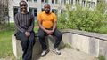 The picture shows Diana Achuch and Obadiah Orang'i sitting on a bench outside a university building. They are both smiling at the camera. The building and some bushes can be seen in the background.