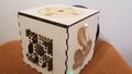 Edge view on a wooden cube. On one side, there is an image of a cobra, on the other a sun wheel, on the third plastic models of Aztec buildings. Not to be seen is the fourth side with a vault opening.
