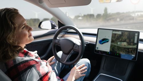 Frau auf dem Fahrersitz eines Autos. Sie berührt das Lenkrad nicht. Neben dem Lenkrad ist ein großes Display, das das Fahrzeug von außen und die Umgebung zeigt.