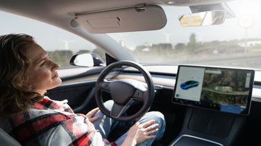 Woman in the driver's seat of a car. She does not touch the steering wheel. Next to the steering wheel is a large display that shows the vehicle from the outside and the surroundings.
