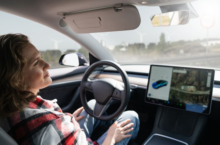 Woman in the driver's seat of a car. She does not touch the steering wheel. Next to the steering wheel is a large display that shows the vehicle from the outside and the surroundings.