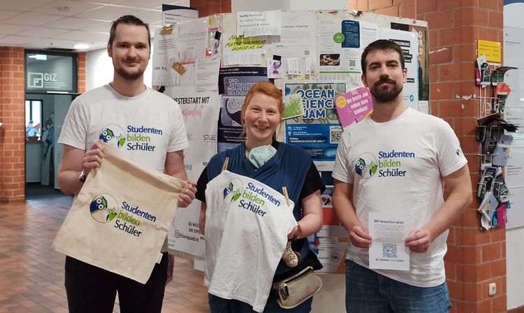 Drei Personen stehen im Mensafoyer vor einem Schwarzen Brett mit vielen Aushängen und halten T-Shirts und Taschen des Vereins hoch.