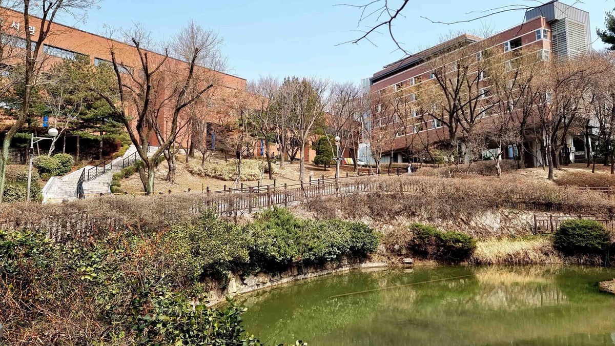 Das Bild zeigt den Campusteich der University of Seoul an einem sonnigen Tag. Im Vordergrund befindet sich ein kleiner Teich mit grüner Vegetation am Ufer. Im Hintergrund erheben sich mehrere moderne Gebäude des Campus, die aus Backstein und Glas bestehen. Zwischen dem Teich und den Gebäuden befindet sich eine leicht abschüssige Grünfläche mit Bäumen und Büschen. Ein kleiner Pfad führt an dem Teich entlang. Der Himmel ist blau und klar.