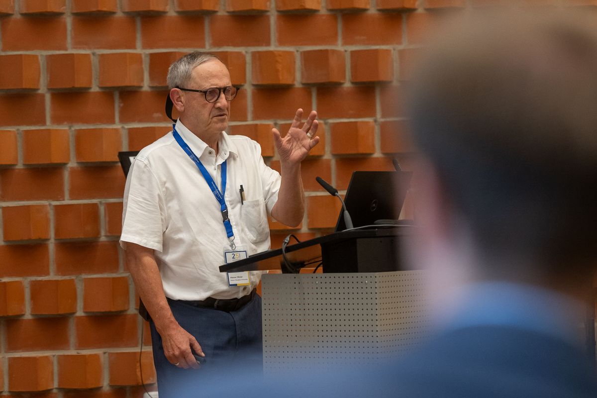 Symposium Ein Mann mit Brille und einem weißen Hemd steht hinter einem Pult und spricht. Er trägt ein Namensschild um den Hals. Er gestikuliert mit seinen Händen, während er spricht. Im Hintergrund ist eine rote Backsteinwand und vor ihm ein verschwommener Teil einer anderen Person zu sehen. Das Bild vermittelt den Eindruck einer Präsentation oder eines Vortrags während des Symposiums.