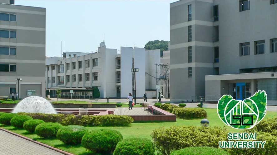 Das Bild zeigt einen Außenbereich des Campus der Sendai University mit mehreren modernen Gebäuden in hellen Farben. Ein gepflegter Garten mit Hecken, Blumenbeeten und einem kleinen Brunnen befindet sich im Vordergrund. Einige Personen sind zu sehen, die sich auf dem Campus bewegen, was eine ruhige und einladende Atmosphäre vermittelt.