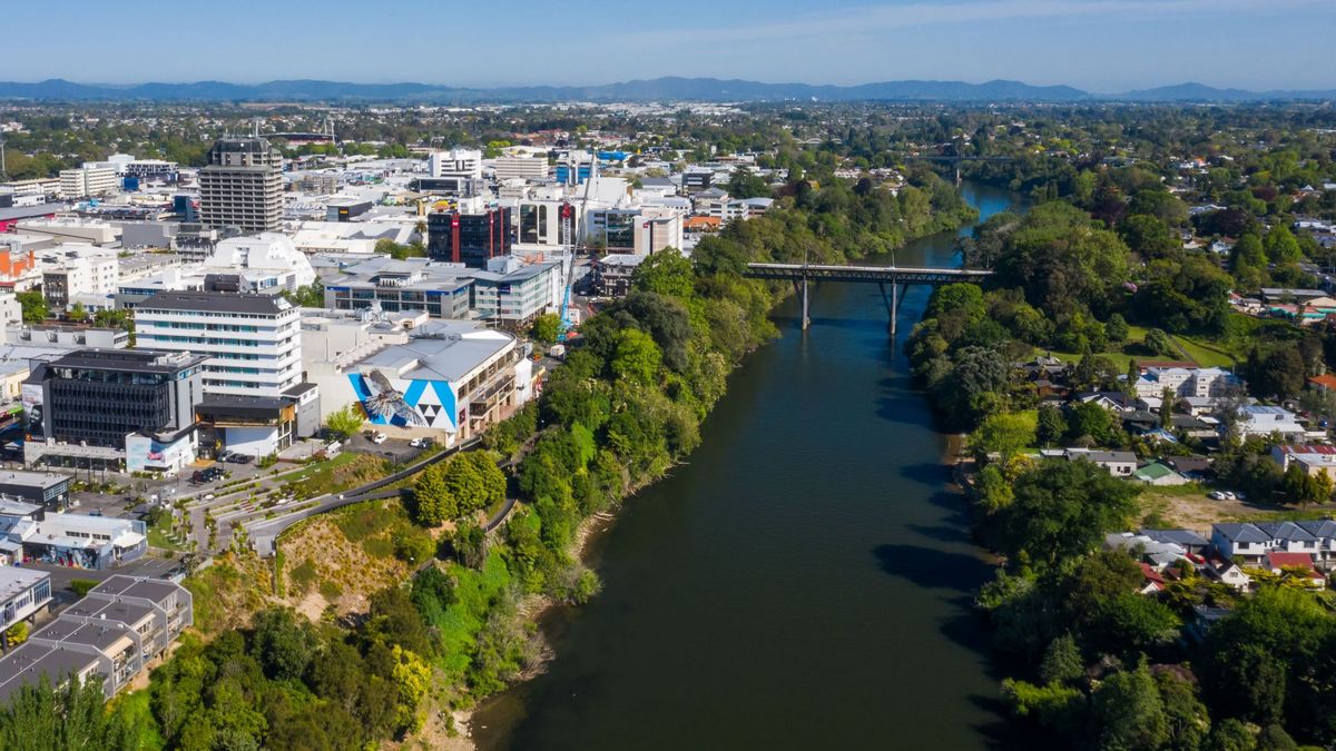 Das Bild zeigt eine Vogelperspektive auf Hamilton, Neuseeland, mit dem Waikato River im Vordergrund. Die Claudelands Bridge überspannt den Fluss und verbindet die Stadtteile miteinander. Moderne Gebäude säumen den Flusslauf und erstrecken sich bis in die Ferne, wo Hügel die Skyline begrenzen.