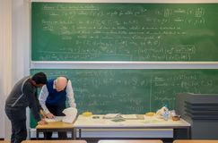 Das Bild zeigt eine große grüne Wandtafel, auf der mit Kreide Formeln geschrieben sind. Vor der Tafel steht ein Tisch, übe den sich in der linken unteren Bildecke zwei Personen beugen und Inhalte eines Papiers besprechen.