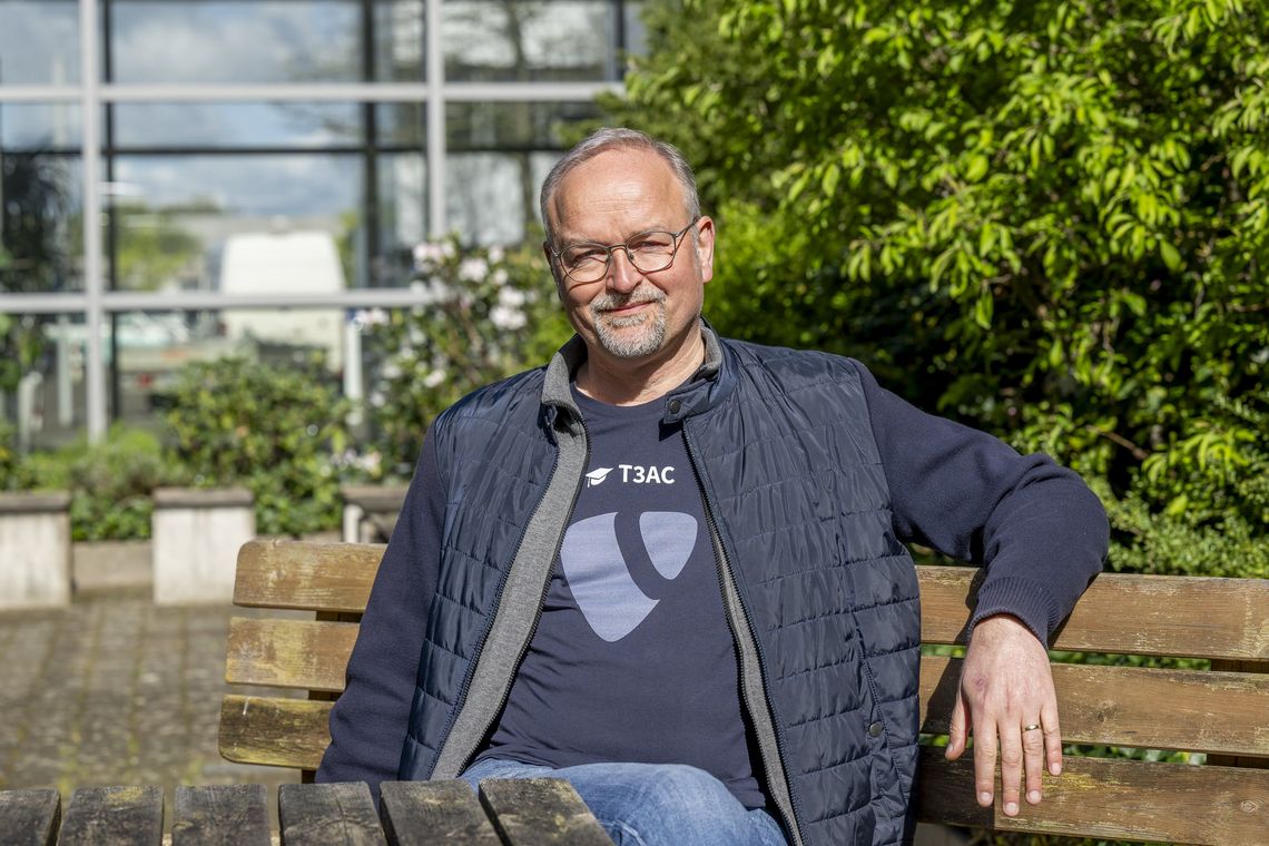 Das Bild zeigt Volker Burggräf. Er sitzt auf einer Bank, hinter ihm ist ein Busch zu erkennen. Er hand einen Arm lässig auf der Bank abgelegt und trägt ein T-Shirt mit dem Logo des Content-Management-Systems Typo3. Er schaut in die Kamera. Die Sonne scheint.