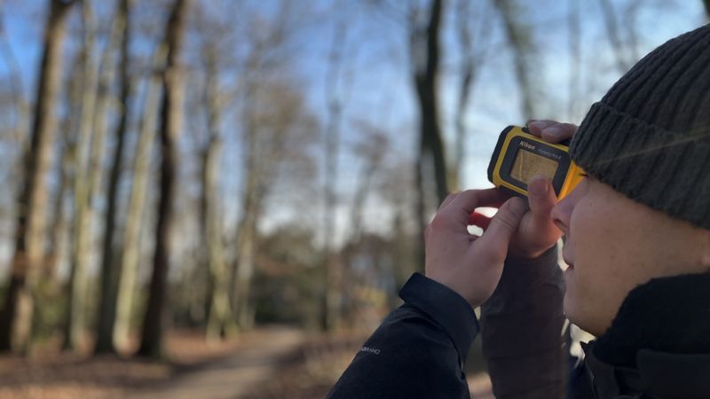 Student hält ein Gerät vor das Auge, im Hintergrund mehrere Bäume.
