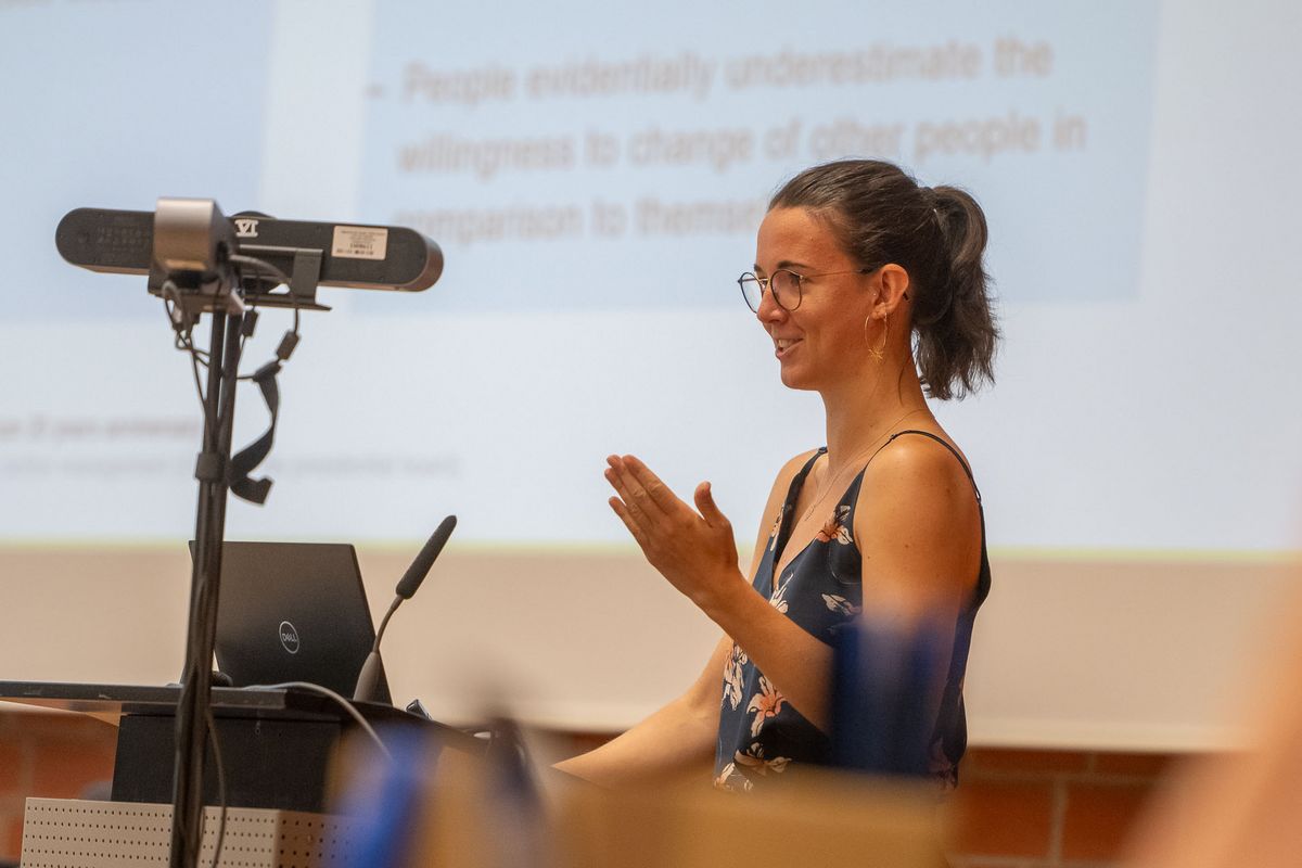 Symposium Eine Frau mit dunklem Haar und Brille steht vor einer Projektion und spricht. Sie trägt ein blaues Top mit Blumenmustern und lächelt, während sie mit ihren Händen gestikuliert. Vor ihr steht ein Laptop und ein Mikrofon auf einem Pult. Der Hintergrund ist verschwommen, aber man kann erkennen, dass sie sich in einem Konferenzraum befindet. Das Bild vermittelt den Eindruck einer Präsentation oder eines Vortrags während des Symposiums.