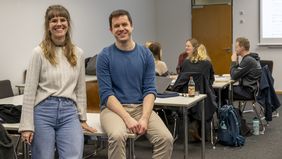 Das Bild zeigt Sophie Berg und Hendrik Wolter. Sie sitzen auf einem Tisch und lächeln in die Kamera. Im Hintergrund sitzt die Gruppe, die der Autor des Textes begleitet hat, beim Spielen zusammen.