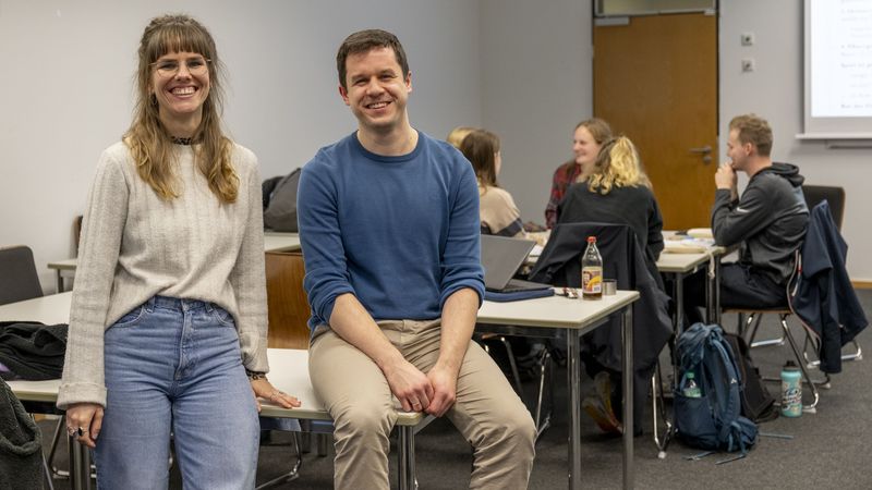 Das Bild zeigt Sophie Berg und Hendrik Wolter. Sie sitzen auf einem Tisch und lächeln in die Kamera. Im Hintergrund sitzt die Gruppe, die der Autor des Textes begleitet hat, beim Spielen zusammen.