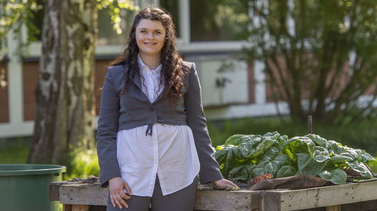 Lillith Rothmund steht im Freien vor Gebäude A 10 im Campusgarten. Sie lehnt sich an ein Hochbeet, in dem Kohl wächst. Sie lächelt in die Kamera.