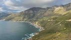 Küste Eine weite Landschaft mit Küstenlinie, Bergen und blauem Meer. Im Hintergrund bewölkte Himmel und grüne Vegetation.