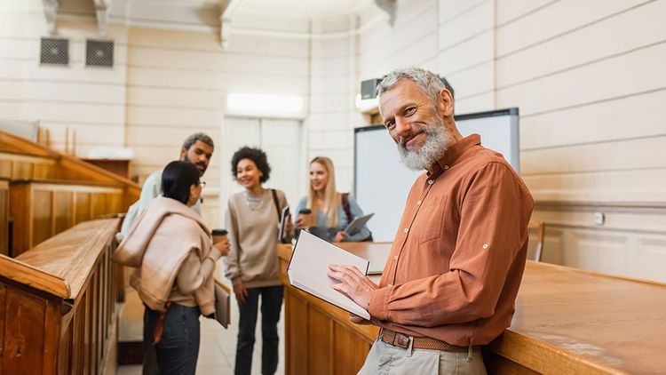 Lächelnder Lehrer, der ein Notebook hält und in die Kamera schaut, in der Nähe von verschwommenen multiethnischen Studenten in der Universität