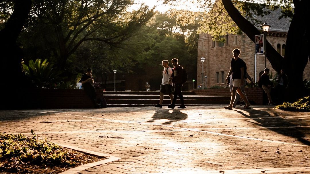 Mehrere Personen gehen auf dem Campus der University of Pretoria über einen gepflasterten Platz. Hohe Bäume werfen lange Schatten, und die Sonne scheint durch das Blätterdach. Im Hintergrund ist ein Gebäude mit hellen Ziegeln zu sehen.