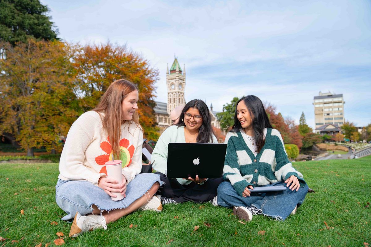 Das Bild zeigt drei Studentinnen, die auf einer grünen Rasenfläche sitzen und gemeinsam an einem Laptop arbeiten. Sie lächeln und scheinen sich über etwas zu unterhalten, was eine positive und kollaborative Lernumgebung suggeriert. Im Hintergrund ist der markante Glockenturm der University of Otago zu sehen, der die Campus-Umgebung verdeutlicht.