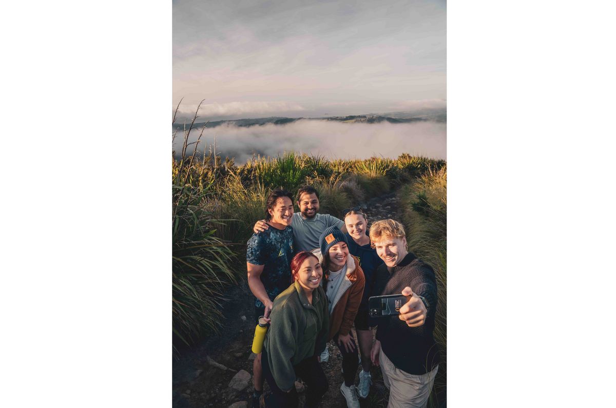 Das Bild zeigt eine Gruppe von Studierenden, die eng zusammensteht und ein Selfie auf einem Hügel mit Blick auf eine nebelverhangene Landschaft macht. Sie lächeln und wirken fröhlich, was auf ein gemeinsames Abenteuer und Freundschaft hindeutet. Die Szene vermittelt ein Gefühl von Gemeinschaft und die Möglichkeit, die natürliche Schönheit der Umgebung von Dunedin zu erkunden.