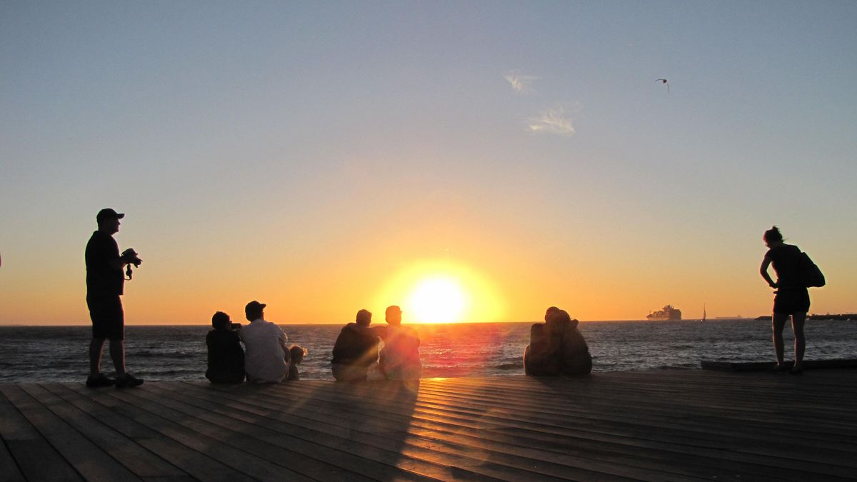 Das Bild zeigt eine Gruppe von Menschen, die einen Sonnenuntergang am Meer beobachten. Die Menschen sind als Silhouetten vor dem hellen, orangefarbenen Himmel zu sehen. Im Hintergrund ist das Meer und ein Schiff zu erkennen. Die Szene vermittelt ein Gefühl von Ruhe und Frieden.