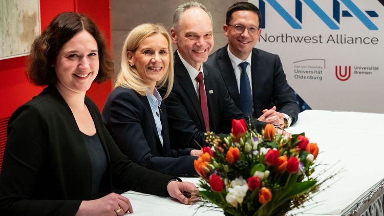 Das Bild zeigt die vier Personen. Sie sitzen an einem Tisch und lächeln in die Kamera. Auf dem Tisch befindet sich eine Vase mit bunten Blumen. Im Hintergrund sind die Logos der Northwest Alliance, der Universität Oldenburg und der Universität Bremen zu erkennen.
