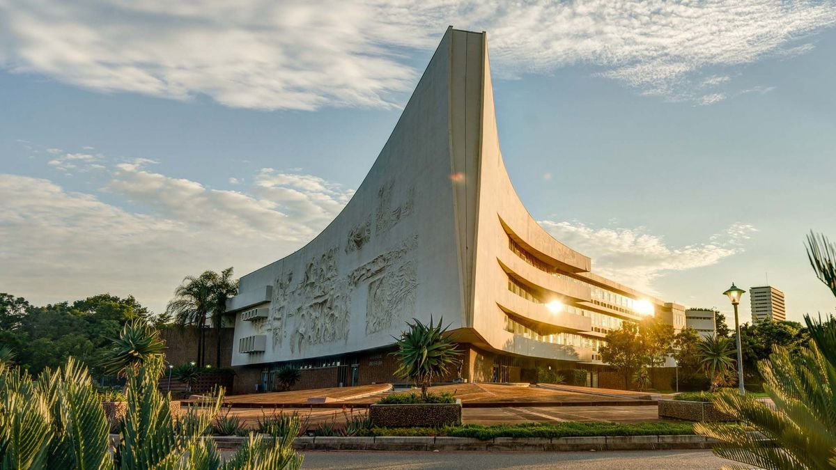  Ein modernes, dreieckiges Gebäude der University of Pretoria ist im goldenen Licht der untergehenden Sonne zu sehen. Die Fassade des Gebäudes ist mit Reliefs verziert und wird von Palmen und Grünflächen umgeben. Im Hintergrund sind weitere Gebäude und der Himmel zu erkennen.