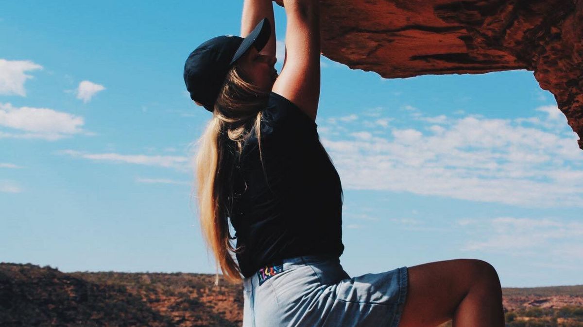 Das Bild zeigt eine Person, die an einem Felsen klettert. Die Person trägt eine Kappe, ein schwarzes T-Shirt und Jeansshorts. Im Hintergrund erstreckt sich eine karge Landschaft mit roten Felsen und trockenem Gelände. Der Himmel ist blau mit einigen Wolken. Die Szene vermittelt ein Gefühl von Abenteuer und sportlicher Betätigung.