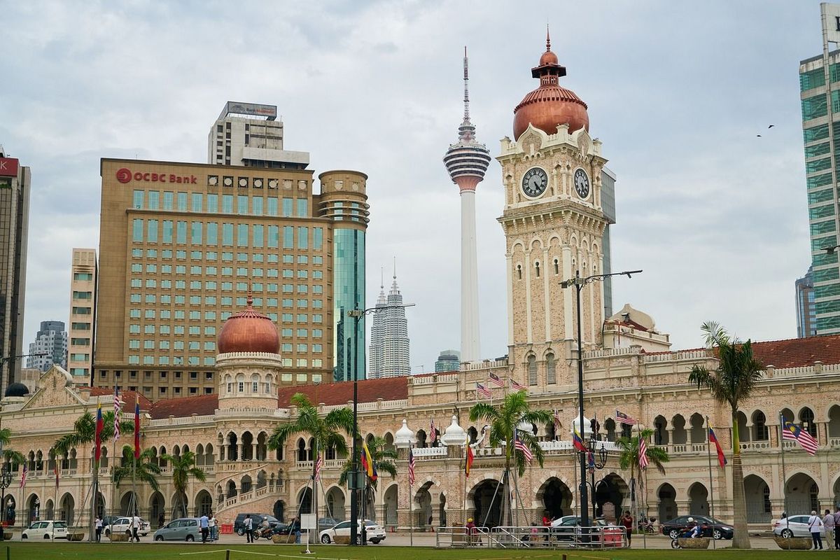 Das Bild zeigt den Sultan Abdul Samad Building und den Kuala Lumpur Tower in Kuala Lumpur, Malaysia. Das Sultan Abdul Samad Building ist ein historisches Gebäude mit Kuppeln und einem Uhrturm. Im Hintergrund ragt der Kuala Lumpur Tower in den Himmel. Vor dem Gebäude befindet sich ein Platz mit Palmen und Flaggen. 