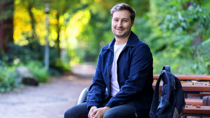 Ein junger Erwachsener sitzt auf einer Bank. Rechts von ihm steht sein Rucksack. Er lächelt in die Kamera.