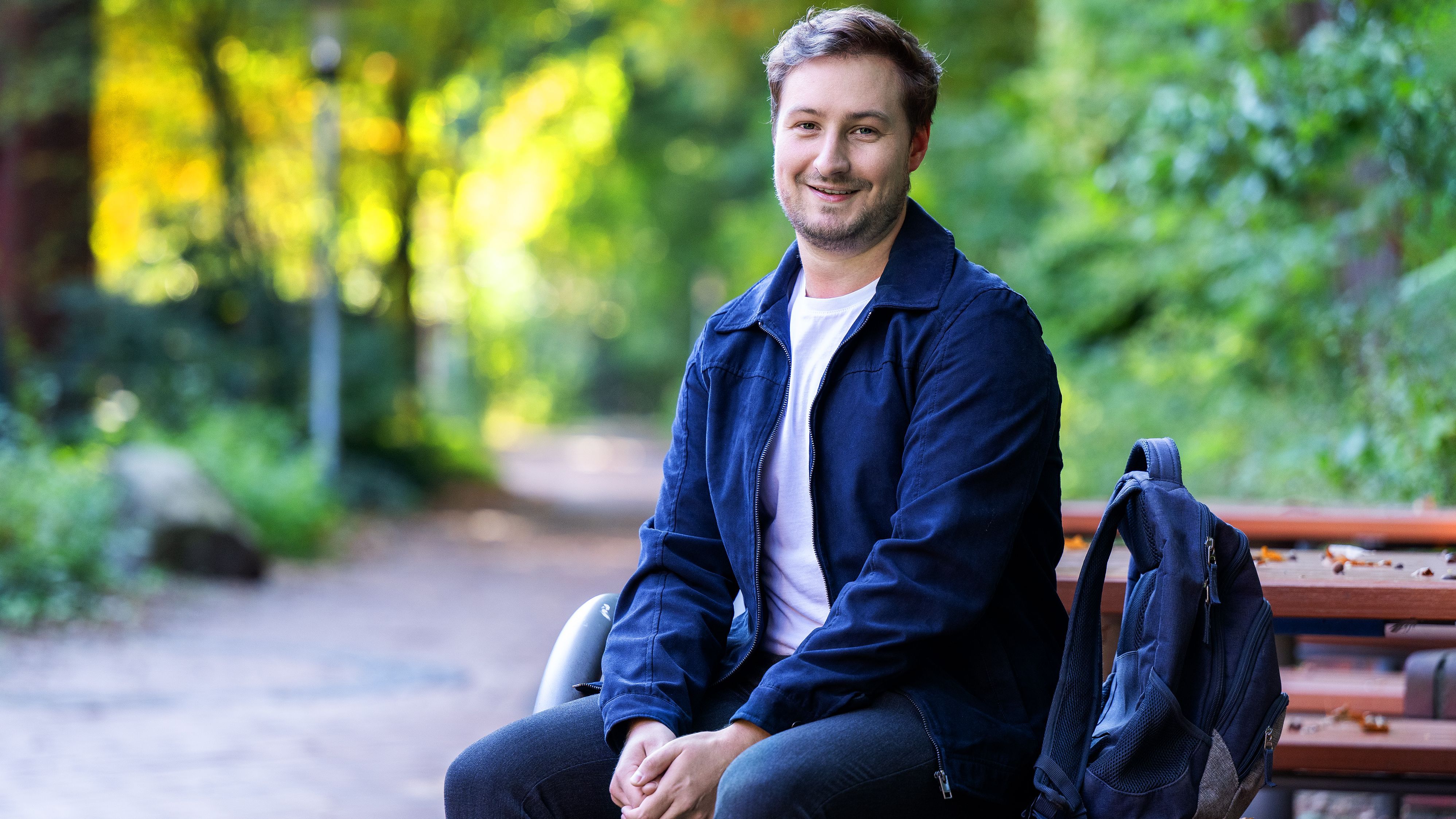 A young adult is sitting on a bench. His rucksack is standing to his right. He is smiling at the camera.
