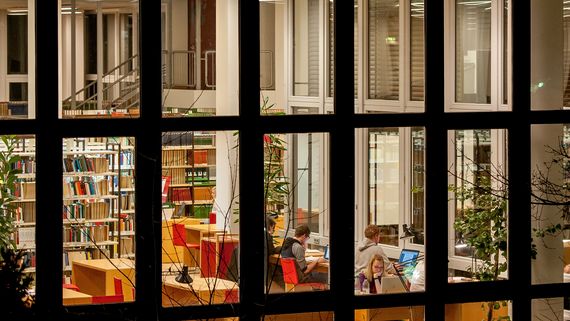 Das Bild zeigt die Bibliothek abends und von außen. In einem Raum kann man Menschen erkennen, die an Schreibtischen arbeiten. im Hintergrund stehen lange Regalreihen mit Büchern.