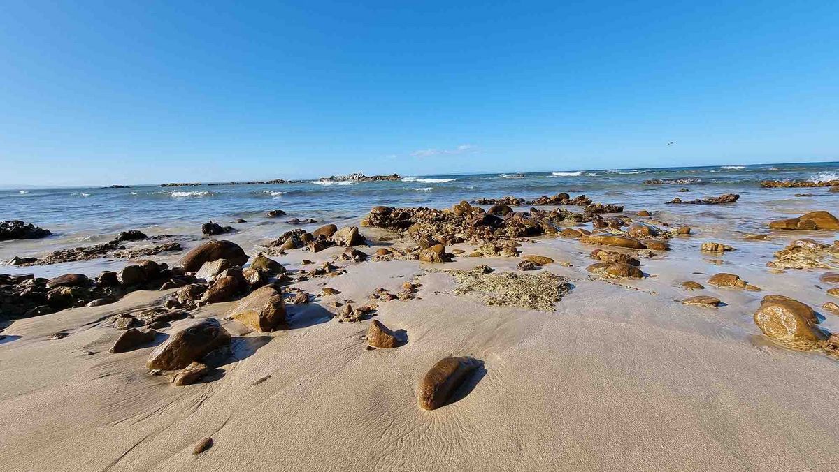 Meer Ein Strand mit felsigen Abschnitten und sanften Wellen. Im Hintergrund das blaue Meer und ein klarer Himmel.