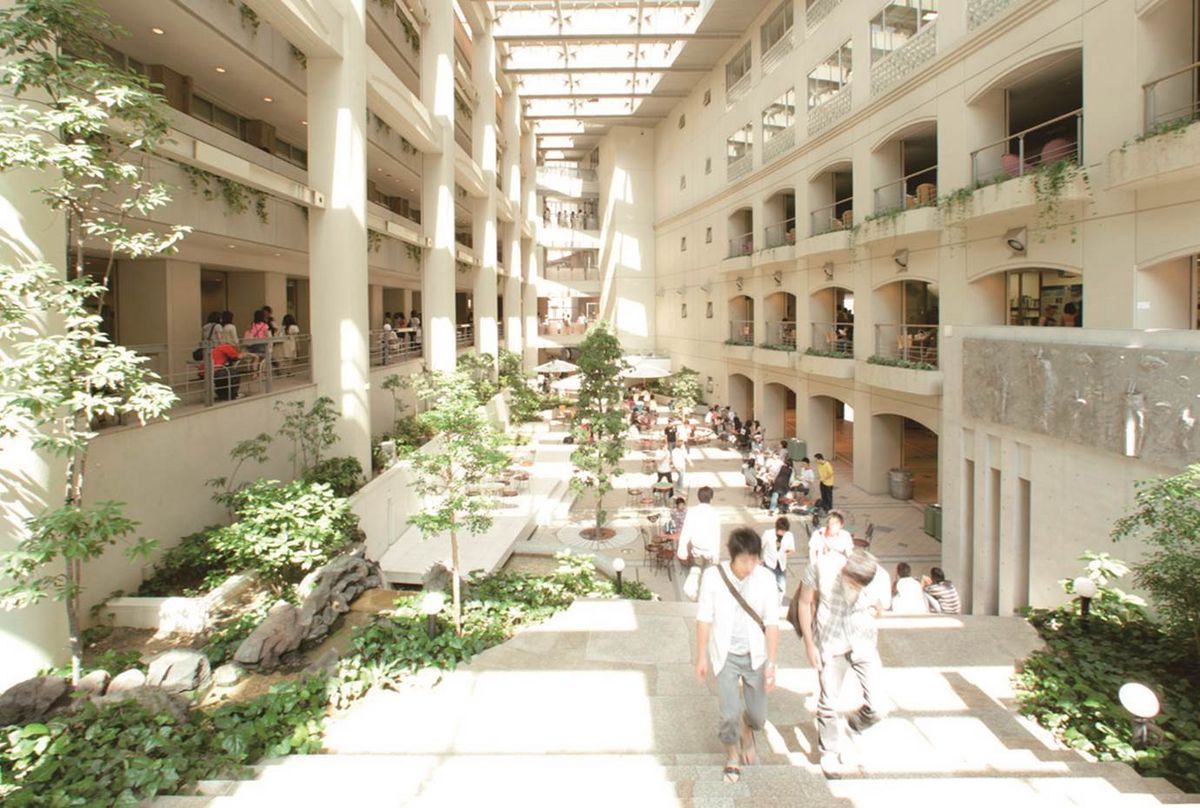 Ein Blick in die Galleria der Chukyo Universität auf dem Nagoya Campus, einen Innenhof mit mehreren Stockwerken, die durch Arkaden verbunden sind. Der Innenhof ist mit Pflanzen, Bäumen und Sitzgelegenheiten gefüllt, und Menschen gehen und sitzen dort. Licht fällt durch das Dach.