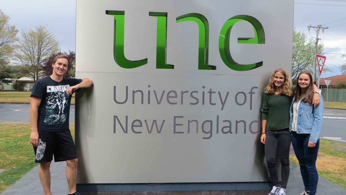 Das Bild zeigt drei lächelnde junge Menschen vor einem großen grauen Schild mit dem Logo der University of New England (UNE). Das Logo besteht aus den Buchstaben "une" in grüner Farbe und dem Schriftzug "University of New England" darunter. Im Hintergrund sind Bäume, eine Straße und Gebäude zu sehen.