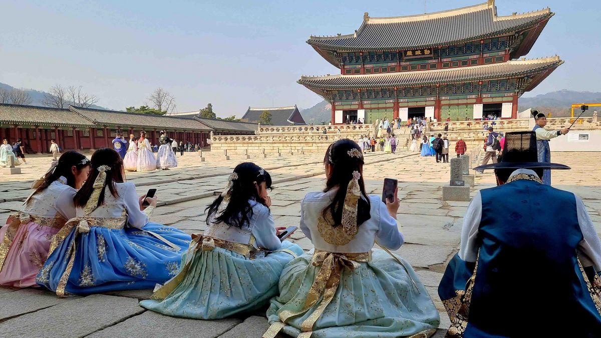 Das Bild zeigt eine Gruppe von Menschen in traditionellen koreanischen Kleidern, Hanboks, vor dem Gyeongbokgung Palast in Seoul. Im Vordergrund sitzen vier Frauen in farbenfrohen Hanboks auf dem gepflasterten Hof und machen Fotos mit ihren Smartphones. Ein Mann in einem dunkleren Hanbok sitzt neben ihnen. Im Hintergrund ist der Gyeongbokgung Palast mit seinen charakteristischen geschwungenen Dächern und farbigen Verzierungen zu sehen. Es sind weitere Besucher*innen zu erkennen, die den Hof erkunden und Fotos machen.