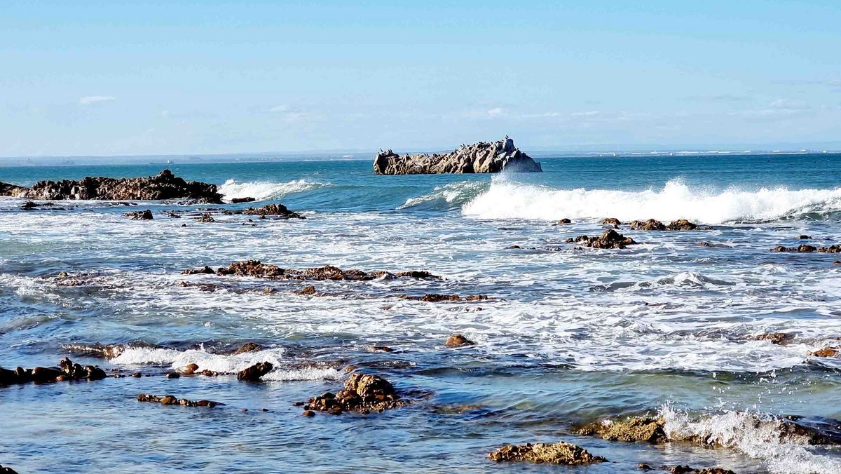 Meer Eine Küstenlandschaft mit felsigen Abschnitten und Wellen. Im Hintergrund eine Insel oder ein Felsen.