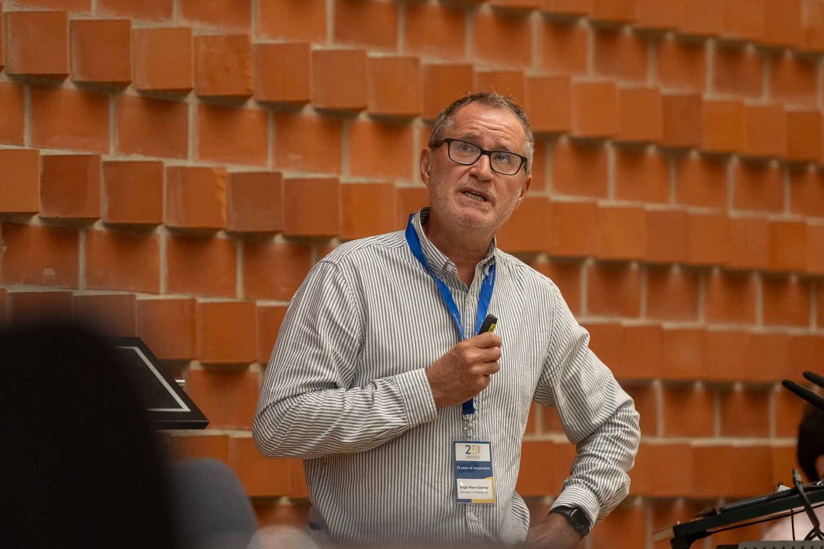 Symposium Ein Mann mit Brille und einem weiß-hellgrau-gestreiften Hemd steht vor einer roten Backsteinwand und spricht. Er trägt eine Lanyard um den Hals mit einem Namensschild. Das Bild vermittelt den Eindruck einer Präsentation oder eines Vortrags während des Symposiums.