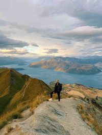 Experience report Gödecke A woman stands on Roys Peak in New Zealand, overlooking a stunning mountain landscape with lakes and valleys. The sky is cloudy, but the sun is shining through.