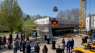 Totale auf den Rohbau, über dem der Richtkranz hängt. Einige Leute stehen um das Gebäude herum.