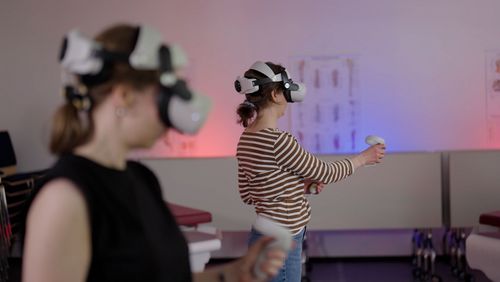 Zwei Personen mit VR-Brille über den Augen stehen in einem Seminarraum und hantieren mit Controllern. 